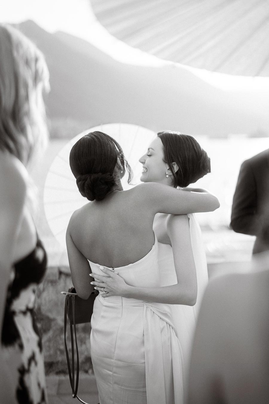 Black and white photo of two women embracing at an outdoor event, with a scenic mountain backdrop and a parasol in the background. One wears an elegant white dress, and the other a strapless gown, both smiling, conveying joy and camaraderie.