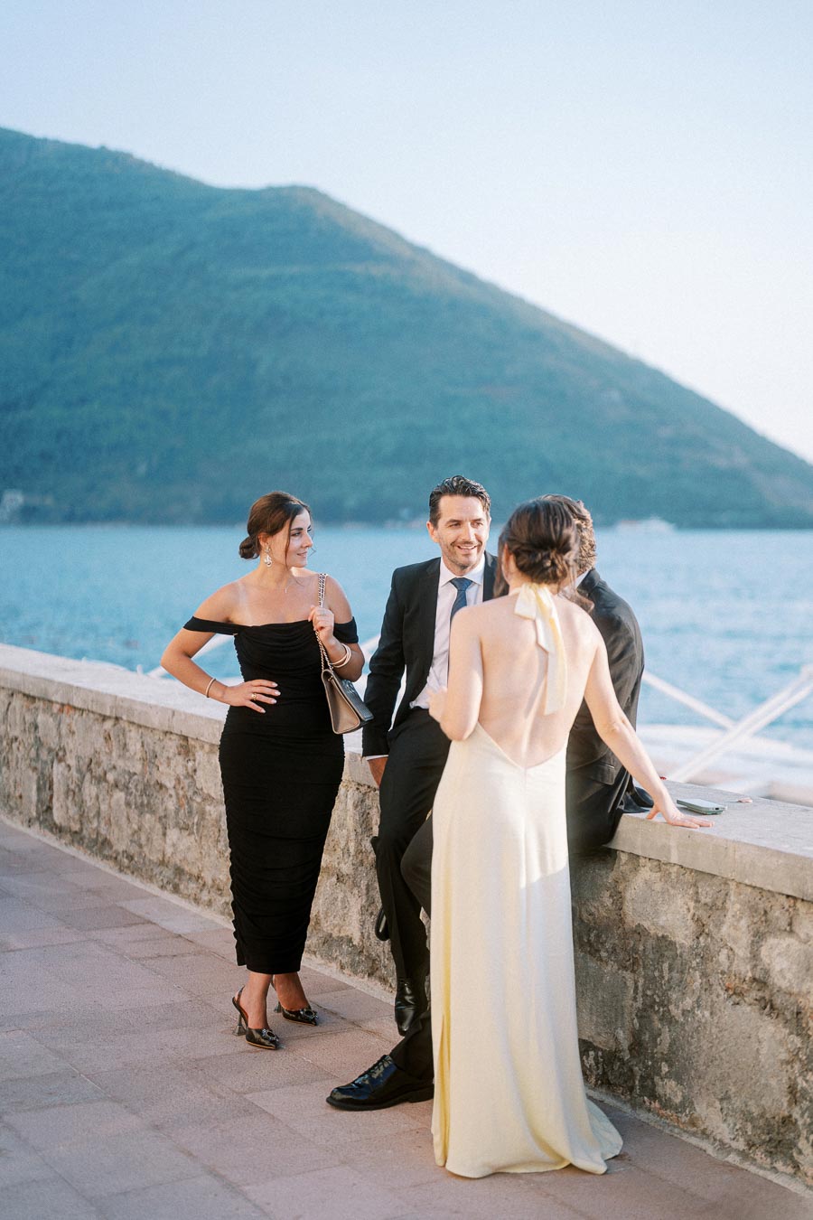 Elegant group of people in formal attire socializing near a scenic waterfront, with a lush mountain backdrop.
