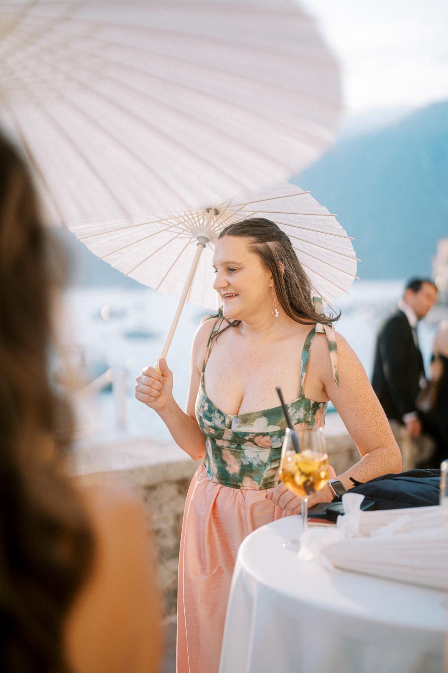 A woman in a floral dress holding a white parasol at an outdoor event by the water, with mountains in the background and a drink on the table.