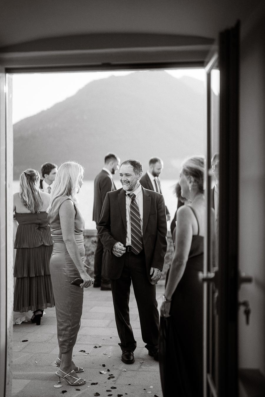 Black and white photo of elegantly dressed people socializing at an outdoor event, with a scenic mountain and lake view in the background.