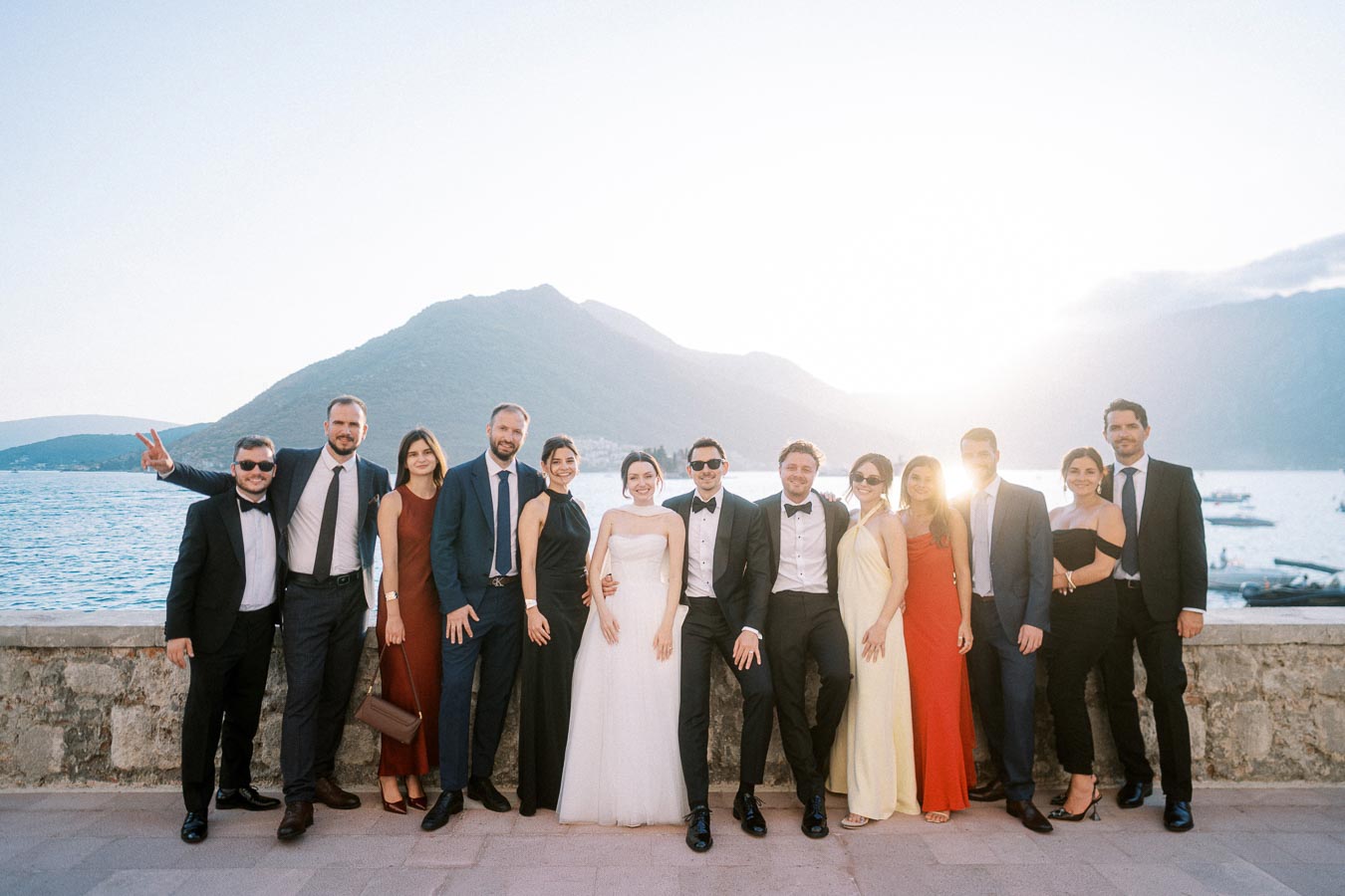 A group of elegantly dressed people posing outdoors at a wedding, with a scenic backdrop of mountains and a waterfront during sunset.