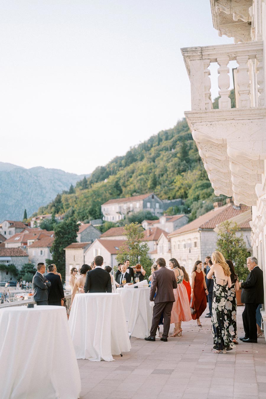 Elegant outdoor wedding reception on a balcony overlooking a scenic landscape with guests socializing around tables, surrounded by historic buildings and lush greenery.