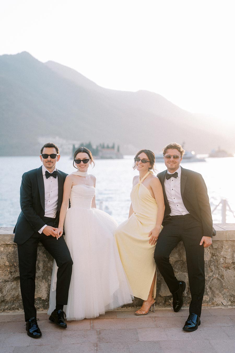 Elegant group at waterfront wedding, featuring two men in tuxedos and two women in stylish dresses, enjoying a scenic backdrop of mountains and water.