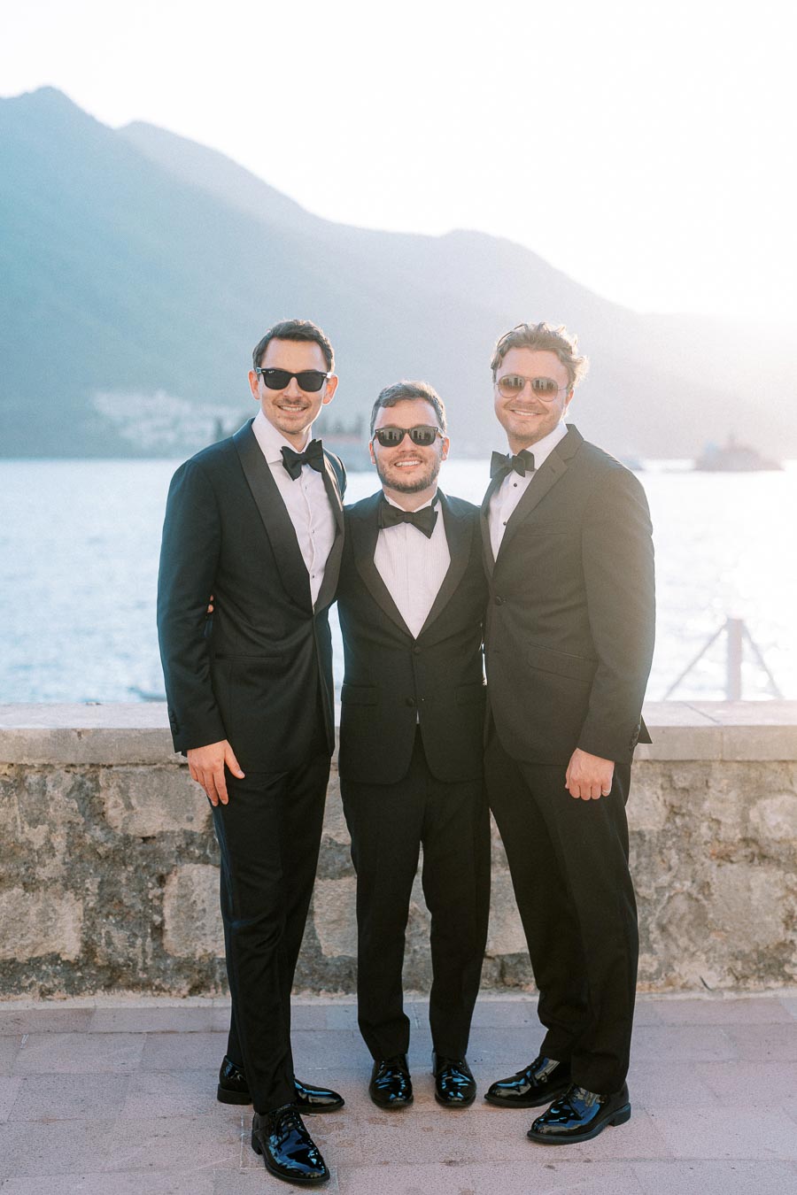 Three men in tuxedos and sunglasses posing for a photograph by a scenic waterfront, with mountains in the background and sunlight highlighting the setting.