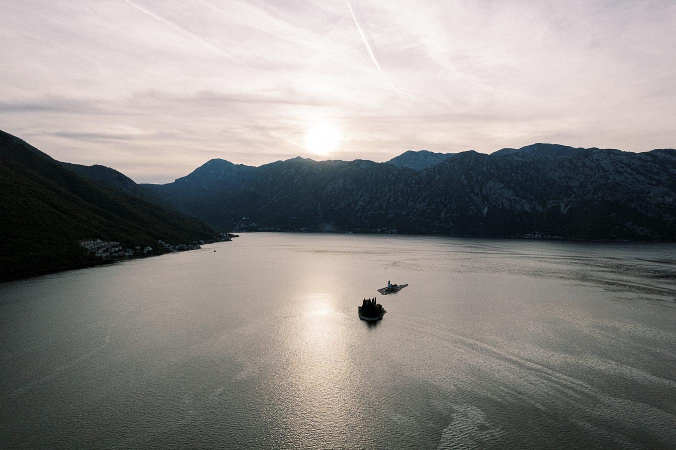Scenic view of a tranquil bay at sunset with a small island and boat, surrounded by majestic mountains in the background.