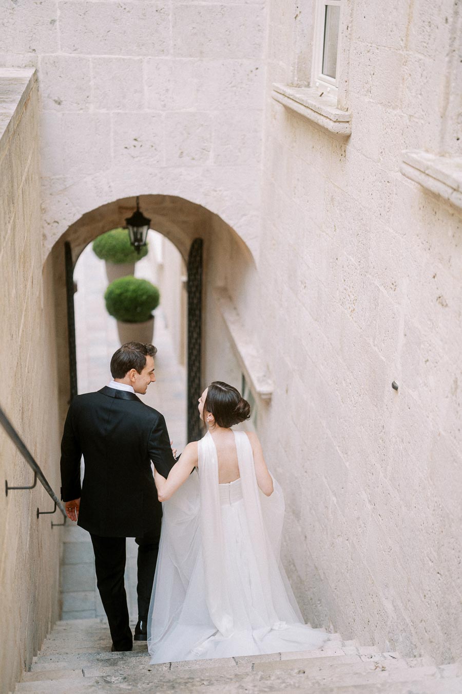 Elegant bride and groom walking down stone staircase in historic building, showcasing romantic wedding moment and architectural beauty.