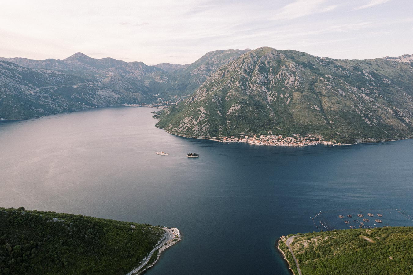 Aerial view of Kotor Bay in Montenegro, showcasing lush green mountains, coastal villages, and tranquil blue waters under a clear sky.