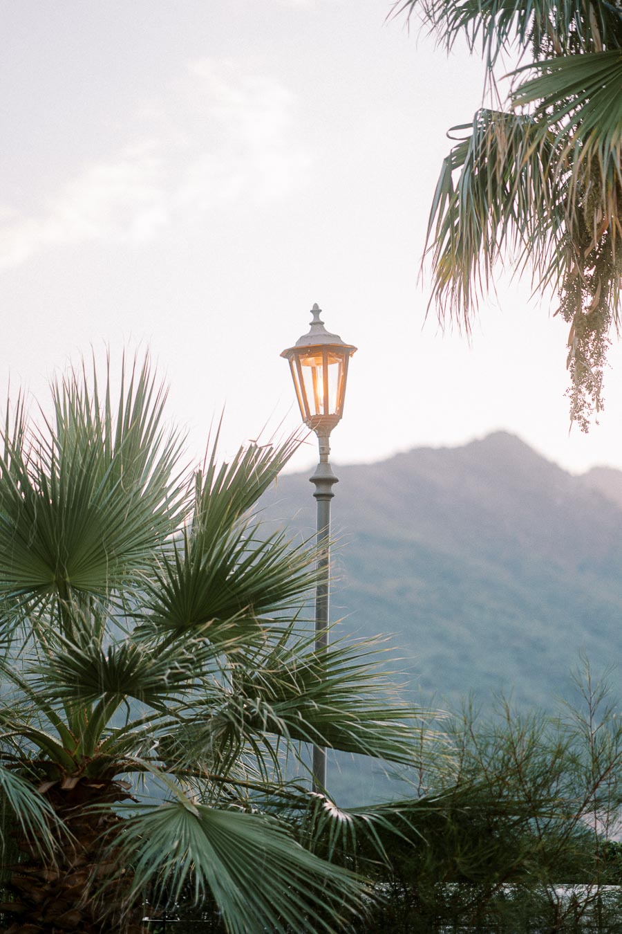 Street lamp illuminating in a serene outdoor setting with palm trees and mountain backdrop at dusk.
