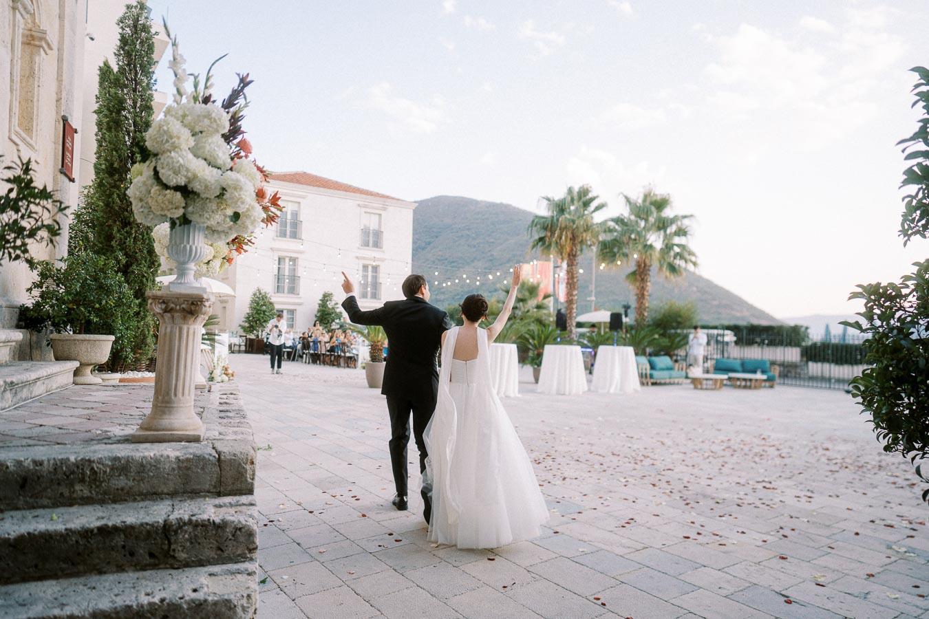 A newlywed couple joyfully walk hand in hand through an elegant outdoor venue decorated with flower arrangements, palm trees, and string lights, against a backdrop of mountains and a clear sky.