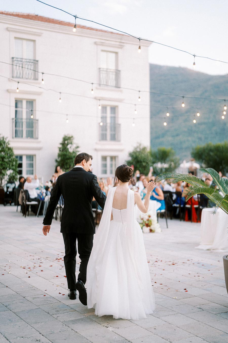 Elegant outdoor wedding reception with a newlywed couple walking hand in hand, surrounded by string lights and guests seated at tables in a picturesque courtyard setting.