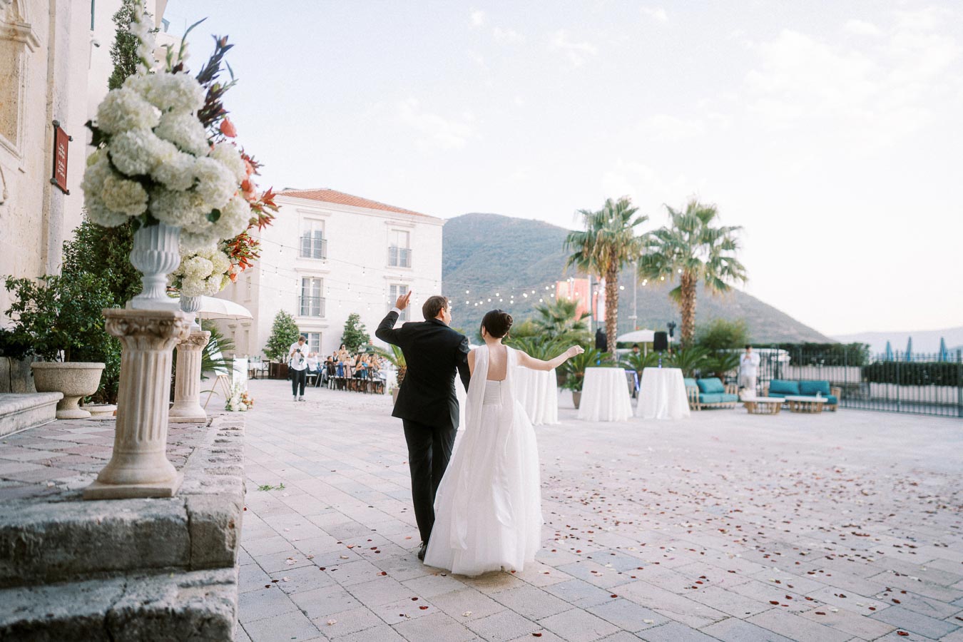 A newlywed couple in elegant attire joyfully walks on a scenic outdoor terrace decorated with floral arrangements, with mountains in the background, during their wedding reception.