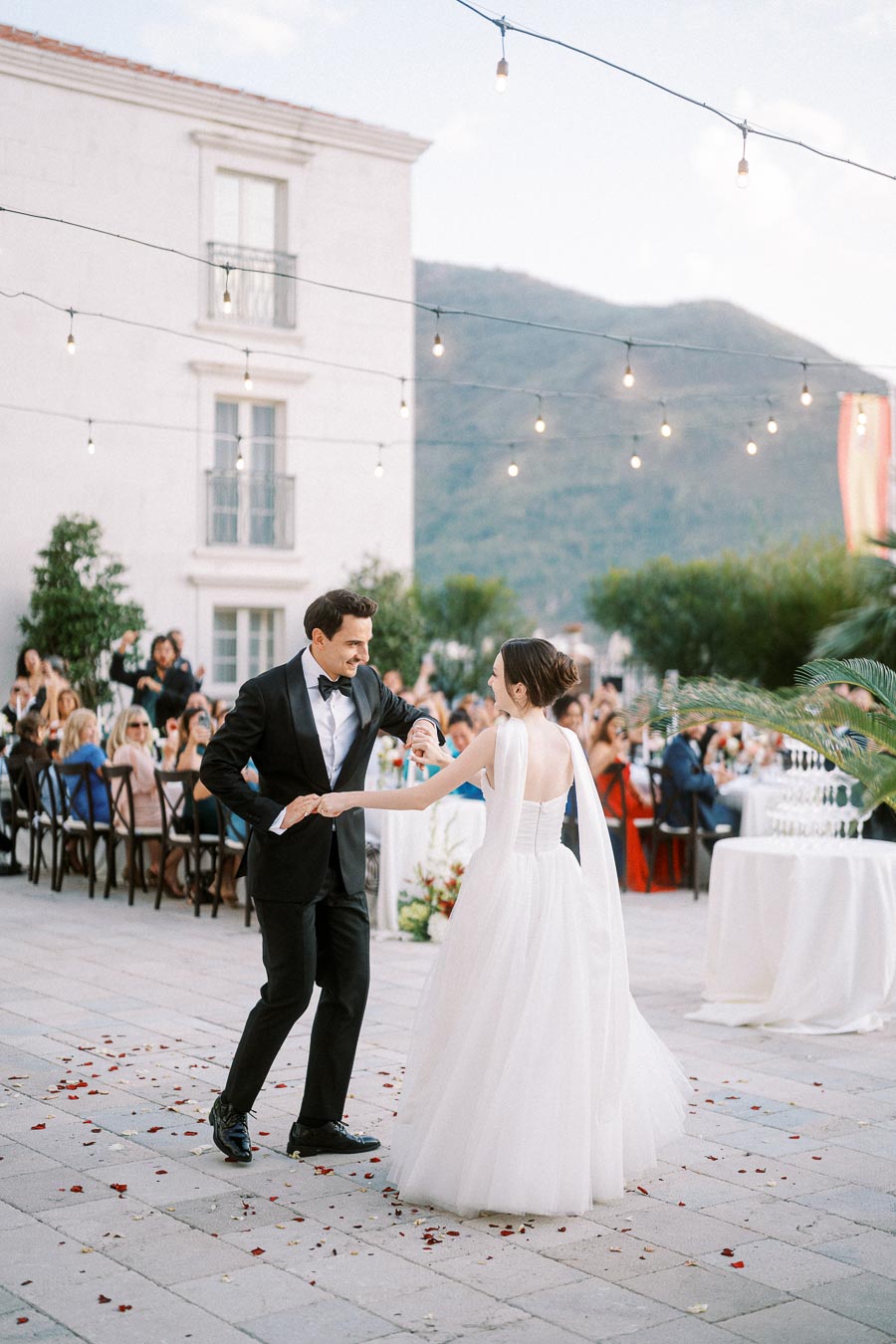 A couple dances joyfully at an elegant outdoor wedding reception, surrounded by seated guests and warm string lights with a scenic mountain backdrop.