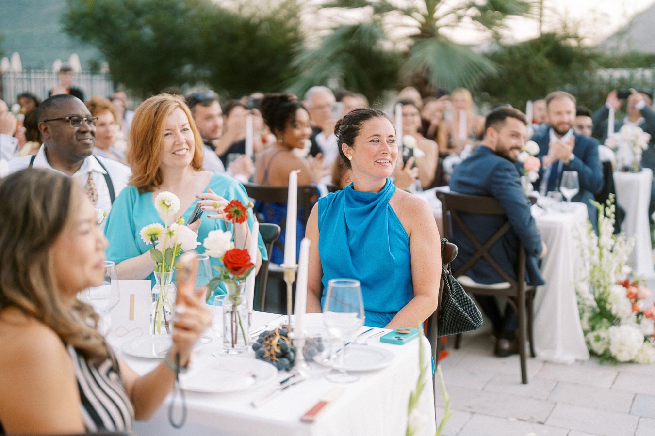 Guests seated at a beautifully decorated outdoor wedding reception, enjoying the event with white tablecloths, flower arrangements, and candles.