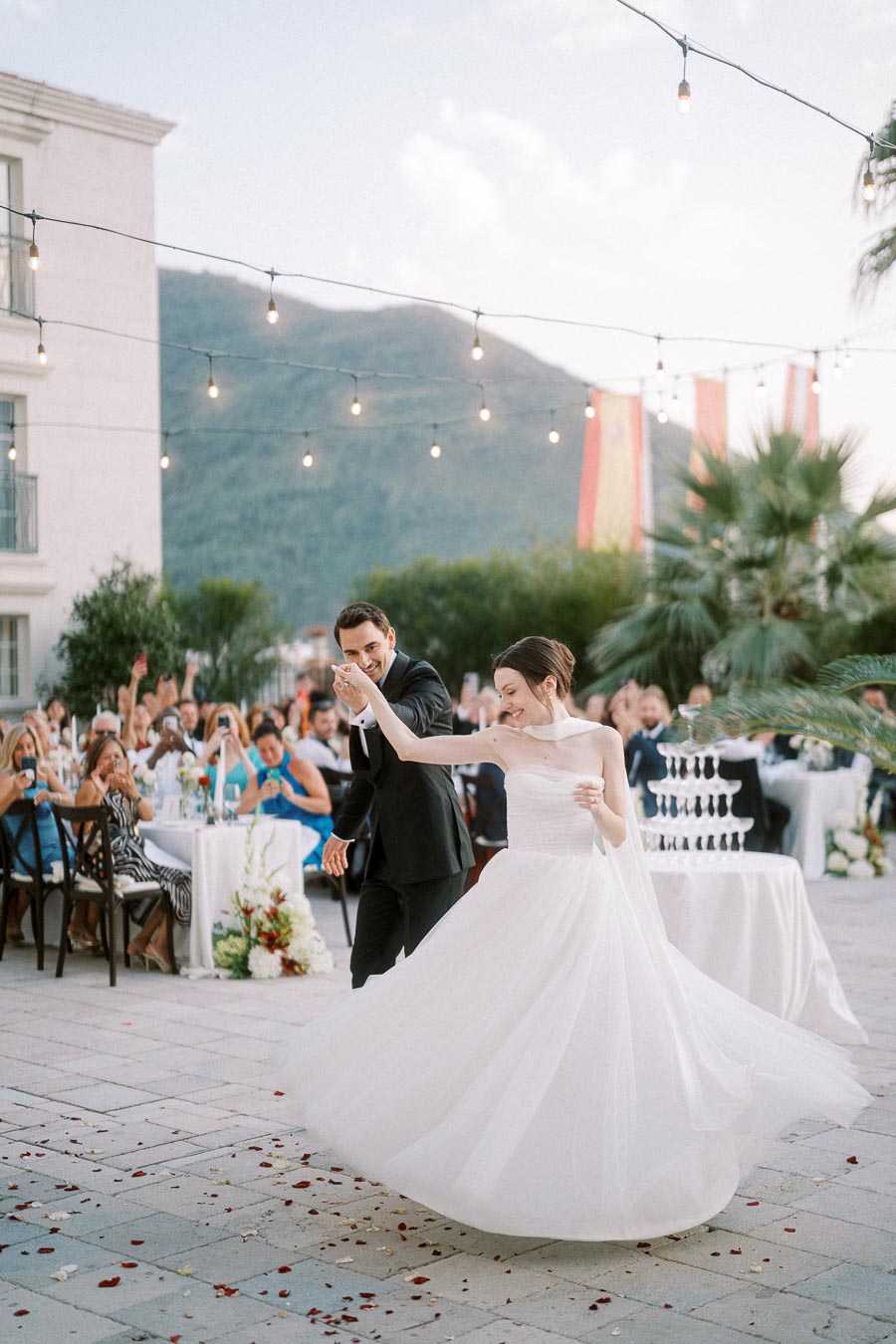 A bride and groom share a joyous first dance at an outdoor wedding reception, surrounded by guests and twinkling string lights. The bride is wearing a flowing white gown, and the groom is in a classic black suit. Elegant tables and a scenic mountain backdrop complete the romantic setting.