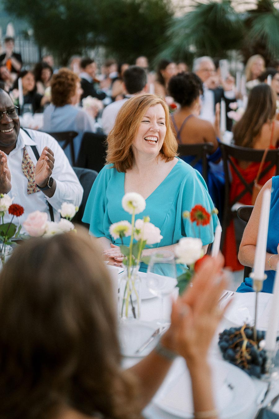 A group of people enjoying an outdoor event, featuring a smiling woman in a turquoise dress surrounded by a lively crowd. The table is decorated with colorful flowers, creating a festive atmosphere.