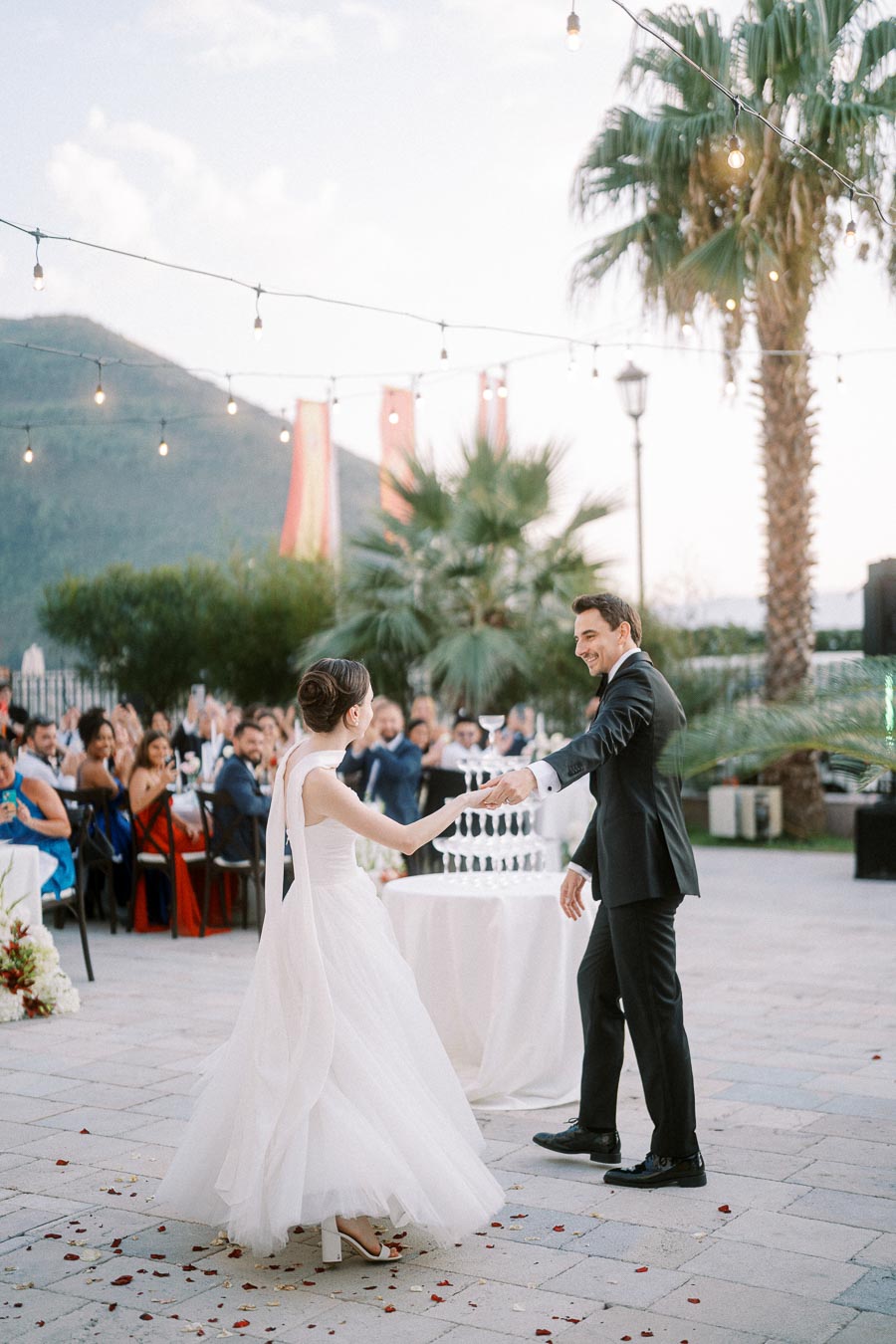 Elegant outdoor wedding dance with a bride in a white gown and a groom in a black suit, surrounded by guests under string lights amidst a scenic mountain and palm backdrop.