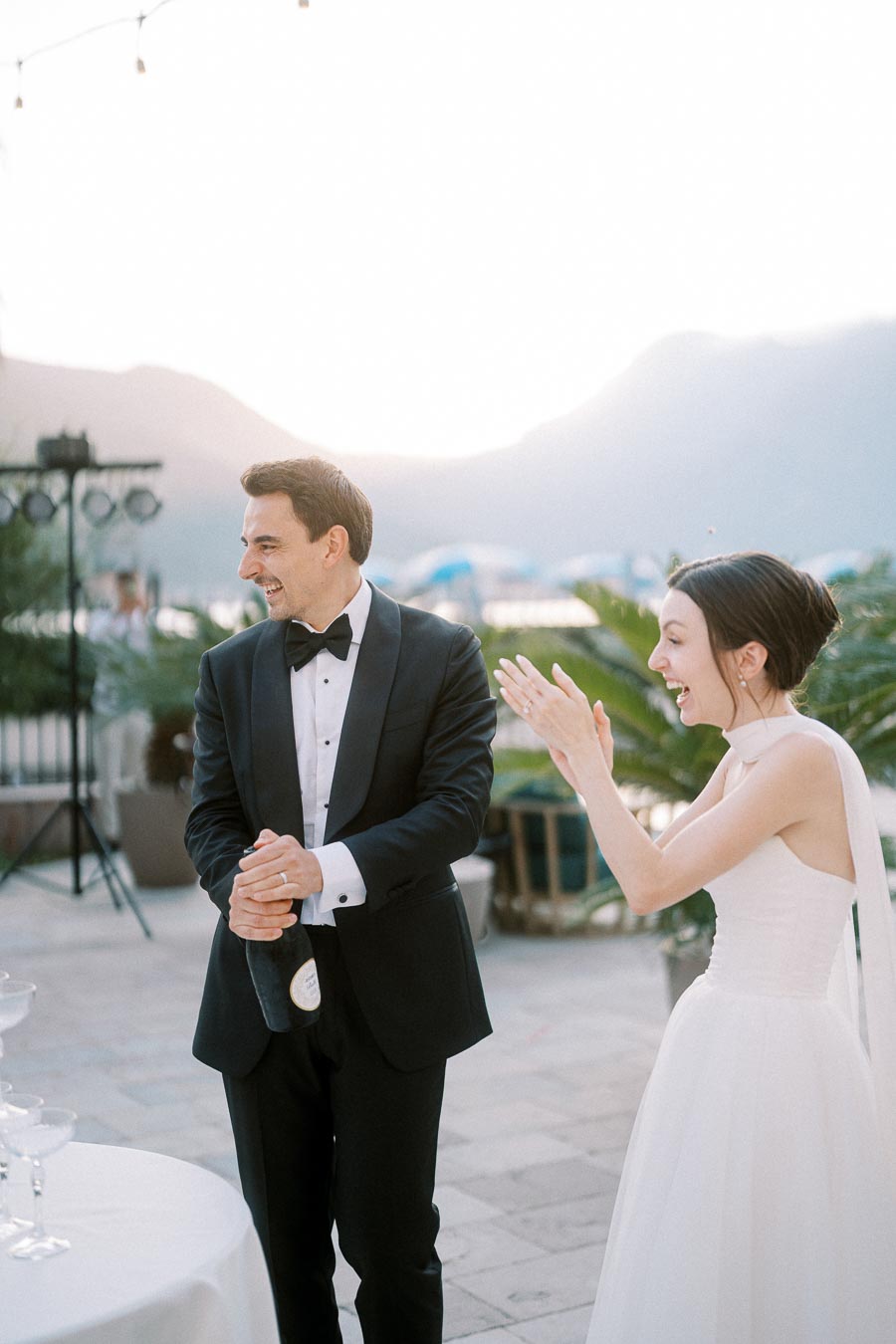 A couple celebrating outdoors at a wedding ceremony, with the groom in a black tuxedo holding a champagne bottle and the bride in a white dress clapping with excitement, set against a scenic mountain backdrop.