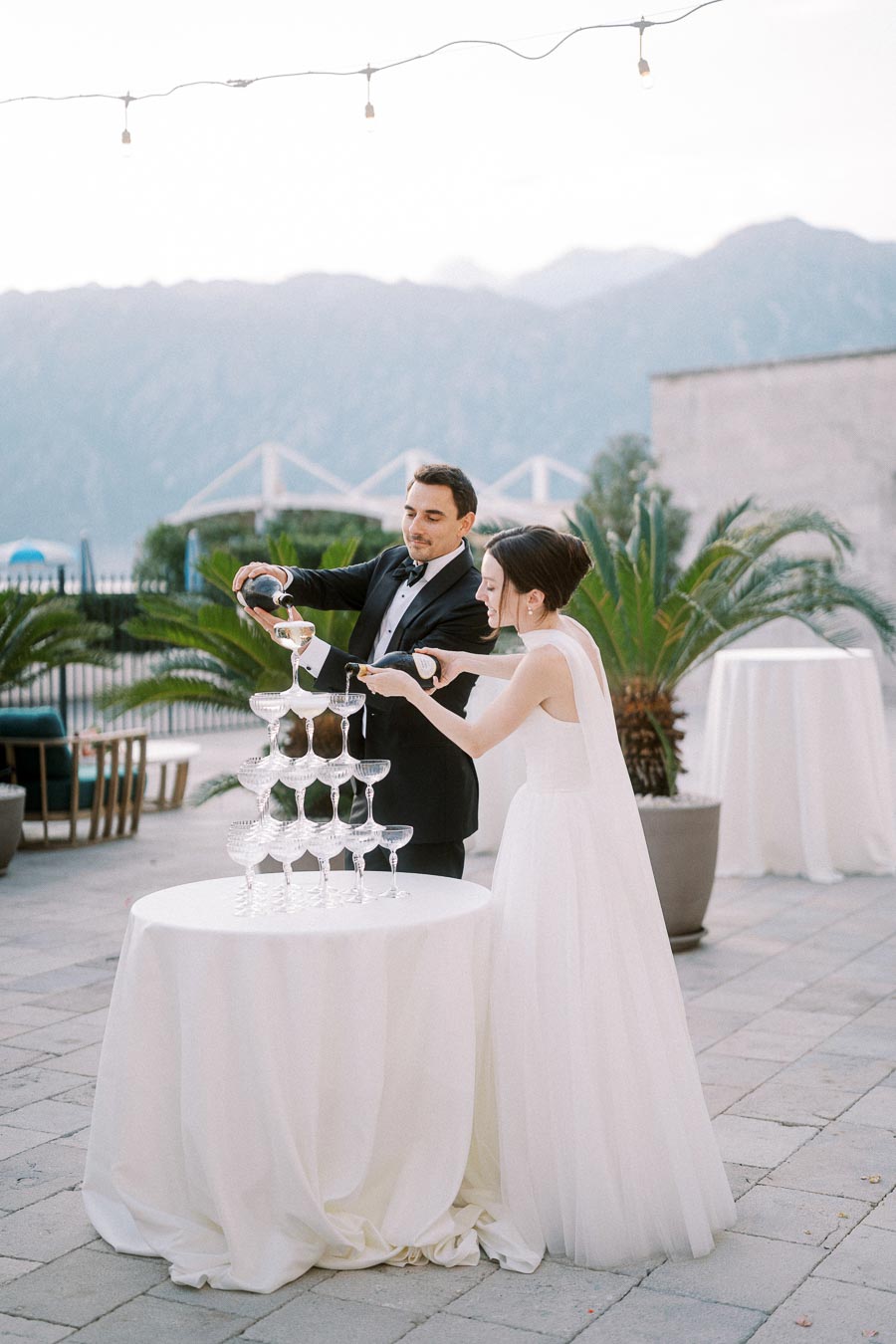A bride and groom pour champagne into a pyramid of glasses at an outdoor wedding reception, with mountains visible in the background.