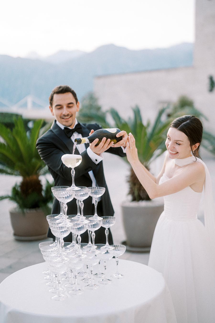 Elegant wedding celebration with a bride and groom pouring champagne into a tiered glass tower outdoors, surrounded by scenic mountains and greenery.