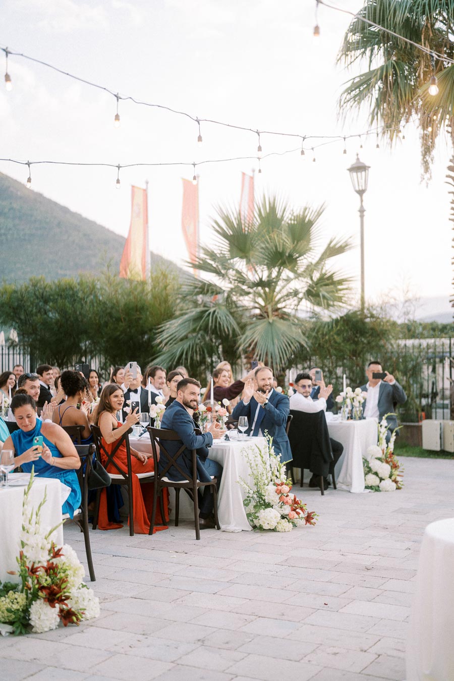 Outdoor wedding reception with guests seated at decorated tables, capturing moments on phones under string lights and palm trees, featuring scenic mountain and garden views.