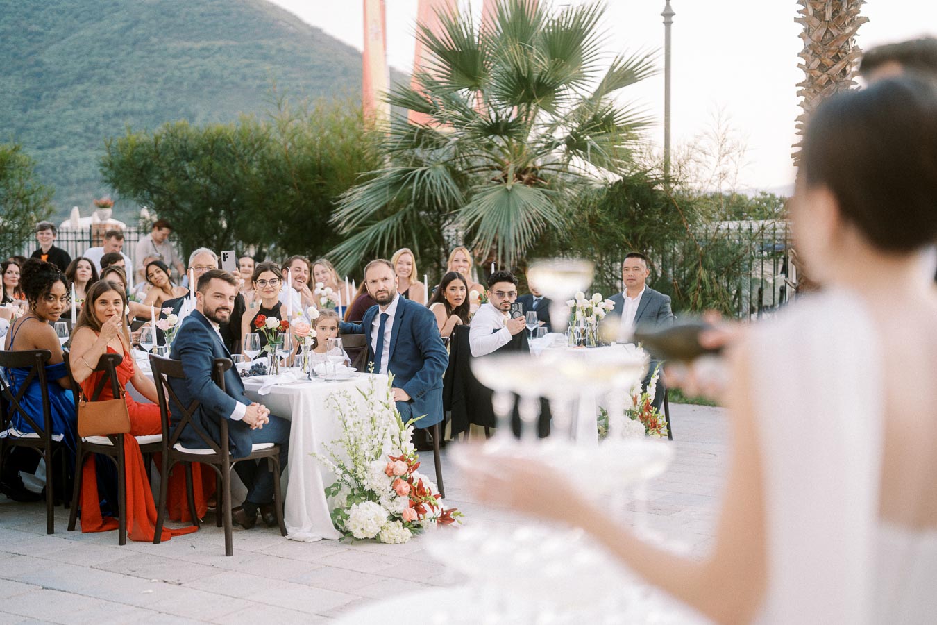 Outdoor wedding reception guests seated at elegant tables with floral centerpieces, focused on a champagne toast under a scenic mountain backdrop.