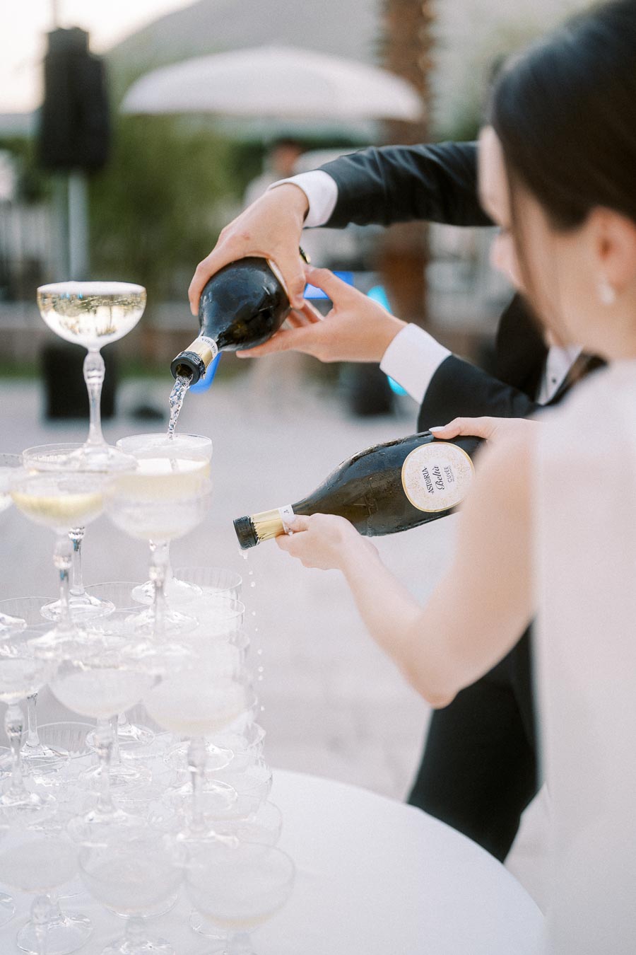Champagne being poured into a cascading tower of elegant glass coupes at an outdoor event, capturing a festive and celebratory atmosphere.