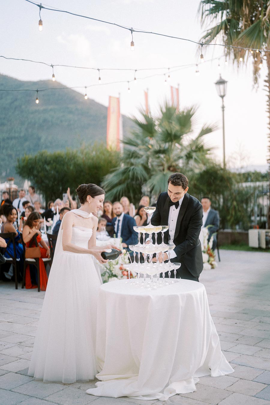 Wedding couple filling a champagne tower at an outdoor reception with guests seated amidst palm trees and string lights, creating a romantic celebration atmosphere.