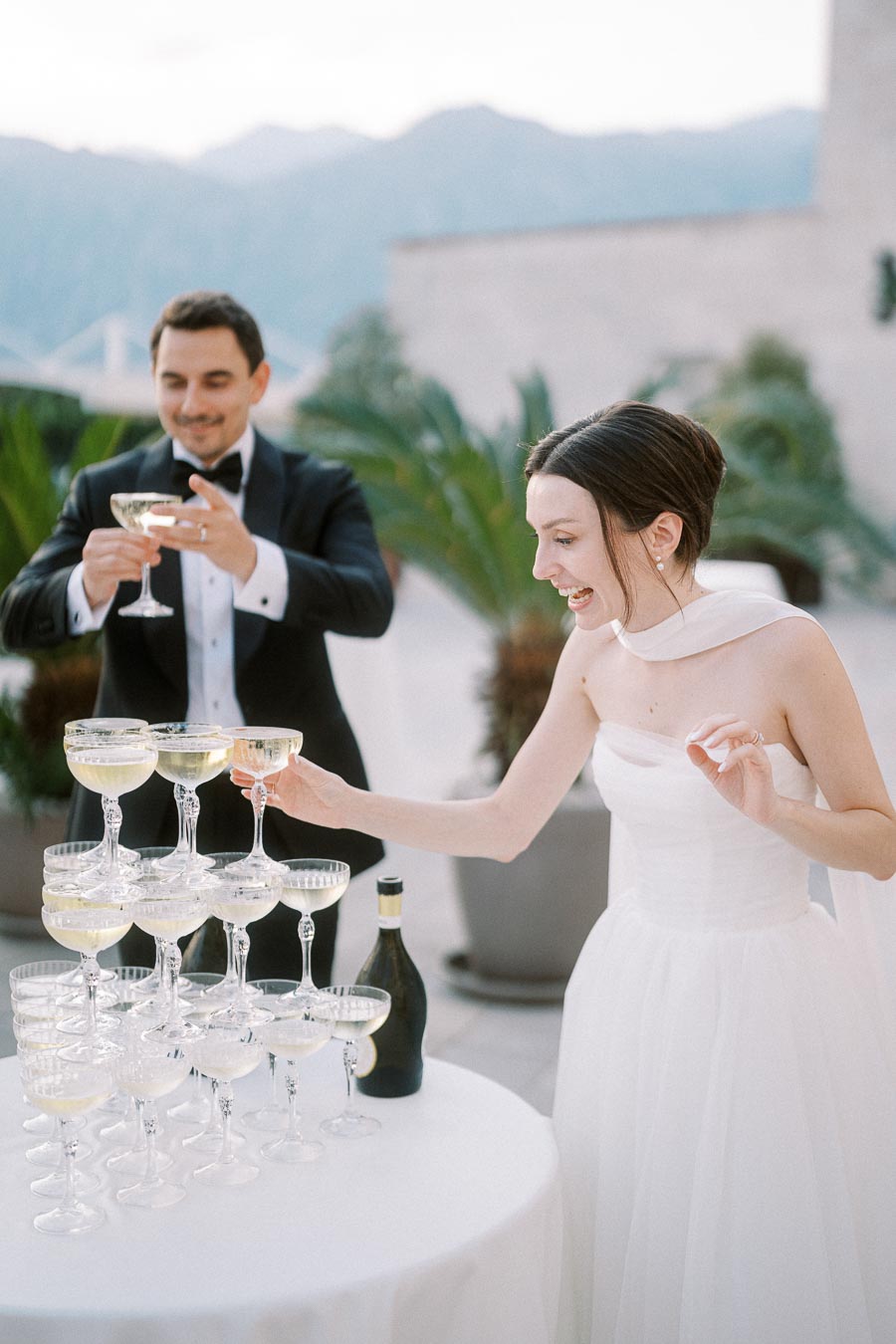 Elegant wedding celebration with bride in white dress pouring champagne into a glass tower, groom in a tuxedo enjoying the moment, and a scenic outdoor backdrop with palm plants.