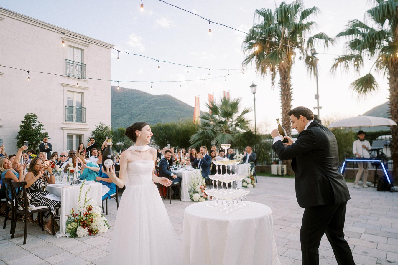 Outdoor wedding reception with bride in white dress and groom in black suit pouring champagne into a tower of glasses. Guests seated at elegantly decorated tables, set against a picturesque backdrop of palm trees and mountains under string lights.