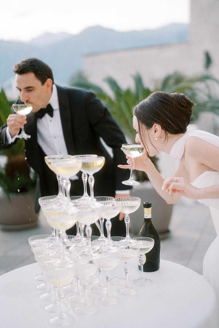 A bride and groom, dressed elegantly at a wedding reception, sip champagne from a pyramid of stacked glasses, surrounded by an elegant outdoor setting.