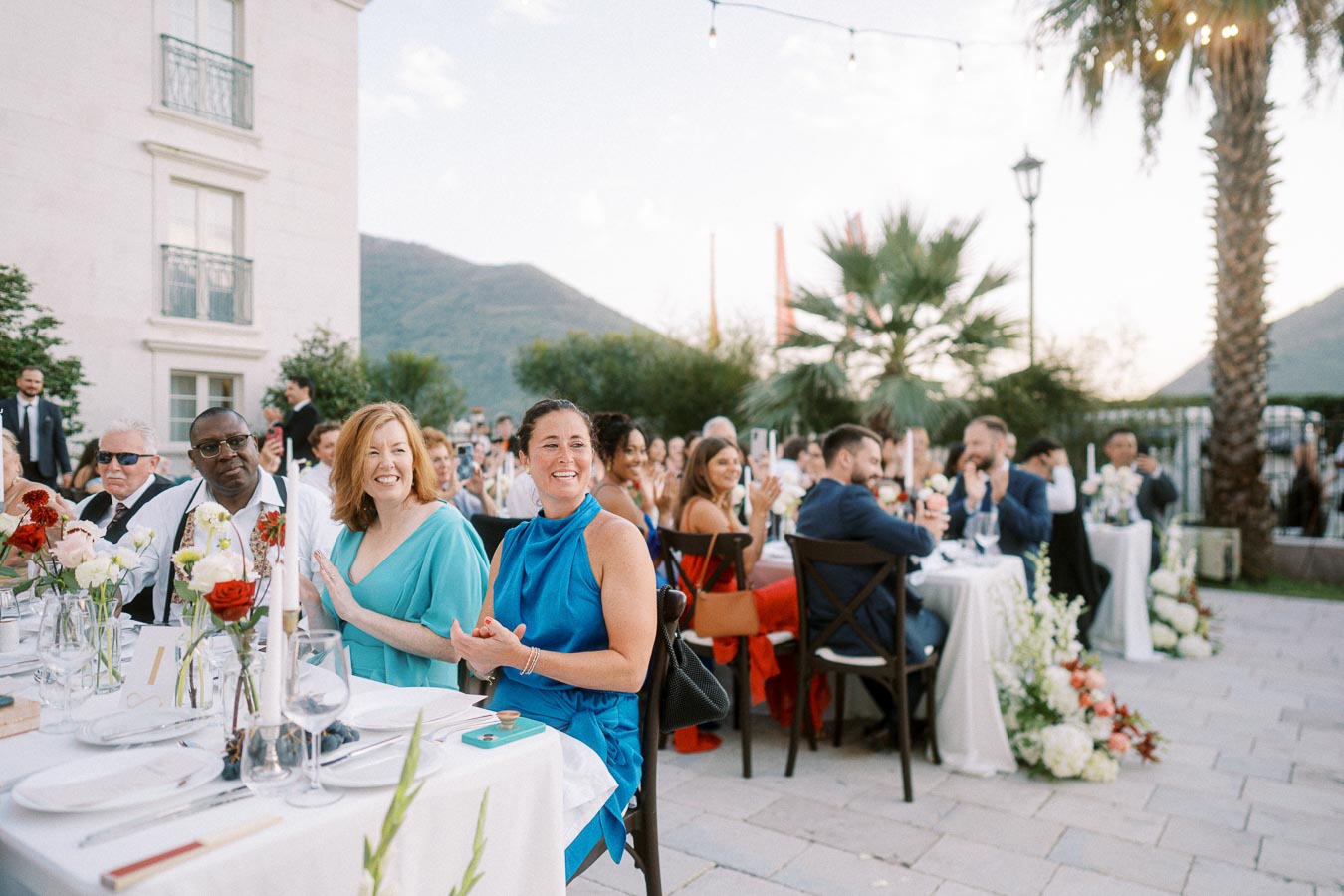 Guests smiling and applauding at an outdoor wedding reception with elegant table settings and floral arrangements, against a backdrop of mountains and palm trees.