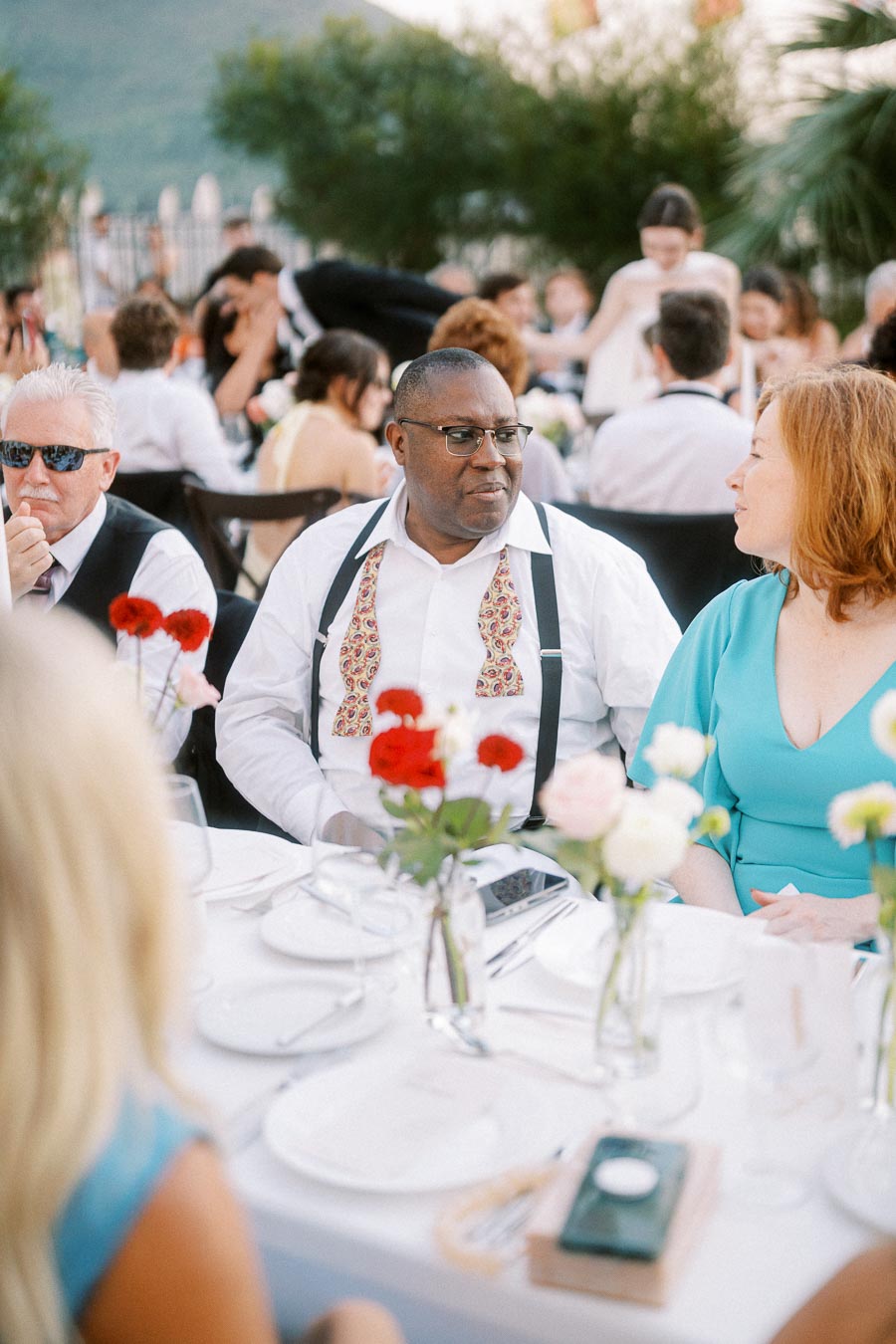 Outdoor wedding reception with guests seated at a table adorned with red and white flowers, engaged in conversation and enjoying the celebration.