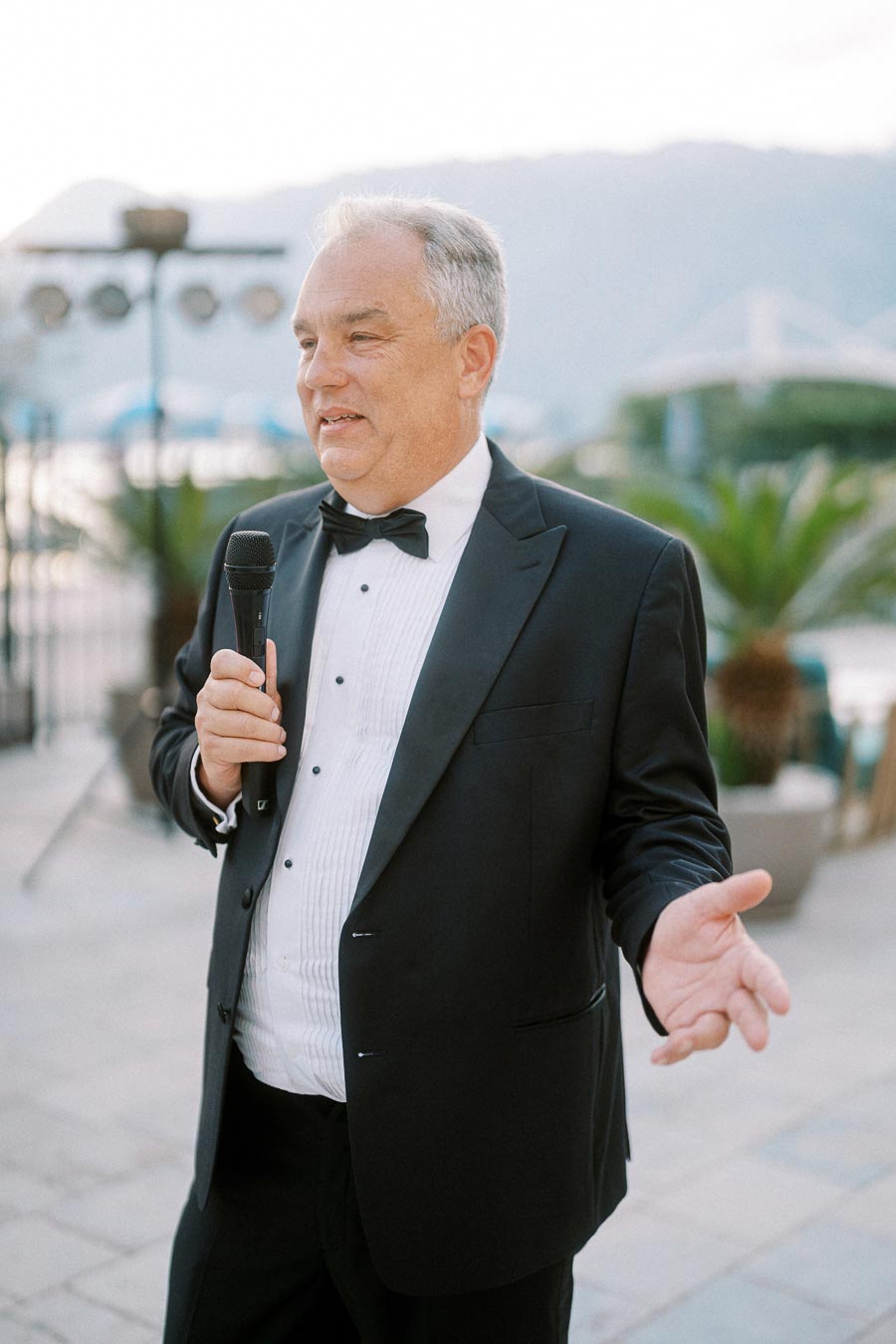 Man in a black tuxedo holding a microphone, speaking outdoors with scenic background of mountains and palm trees.