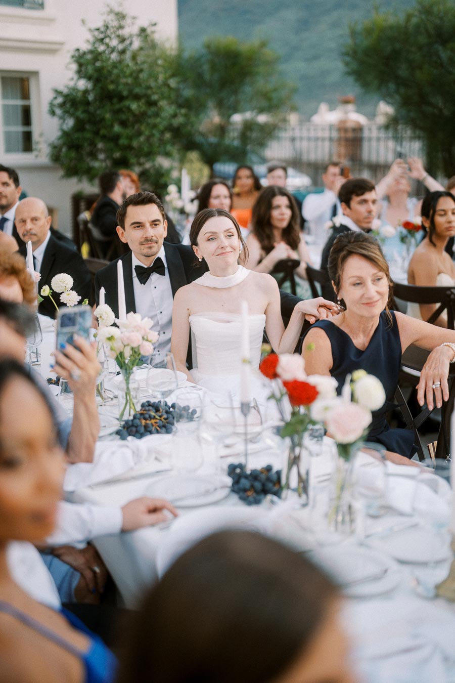Outdoor wedding reception with guests seated at a beautifully decorated table adorned with flowers and candles, featuring a bride in a white dress and a groom in a tuxedo, surrounded by attendees enjoying the celebration.