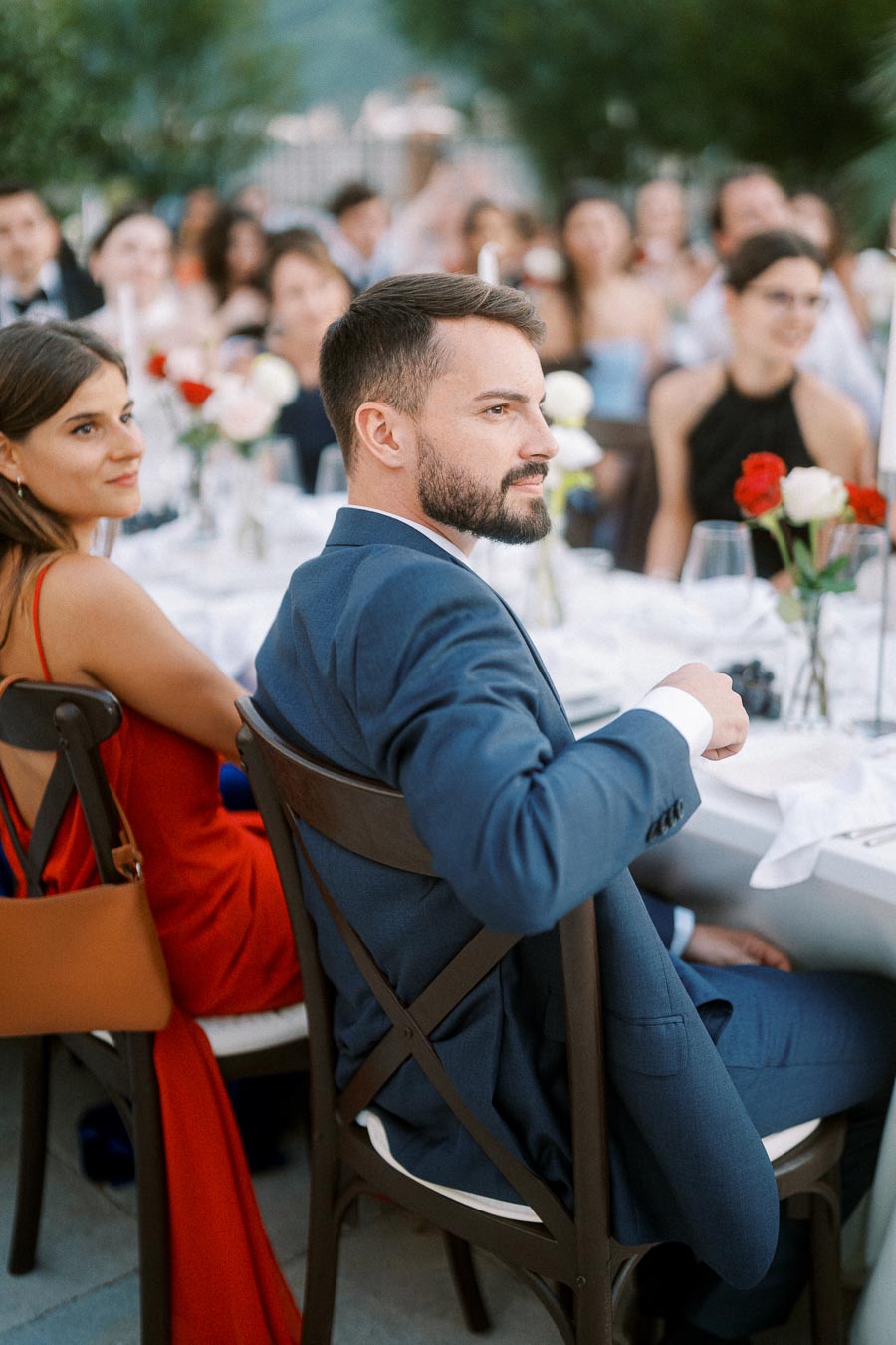 Guests seated at an elegant outdoor event, with a man in a blue suit and a woman in a red dress at a beautifully decorated table adorned with flowers and glassware.