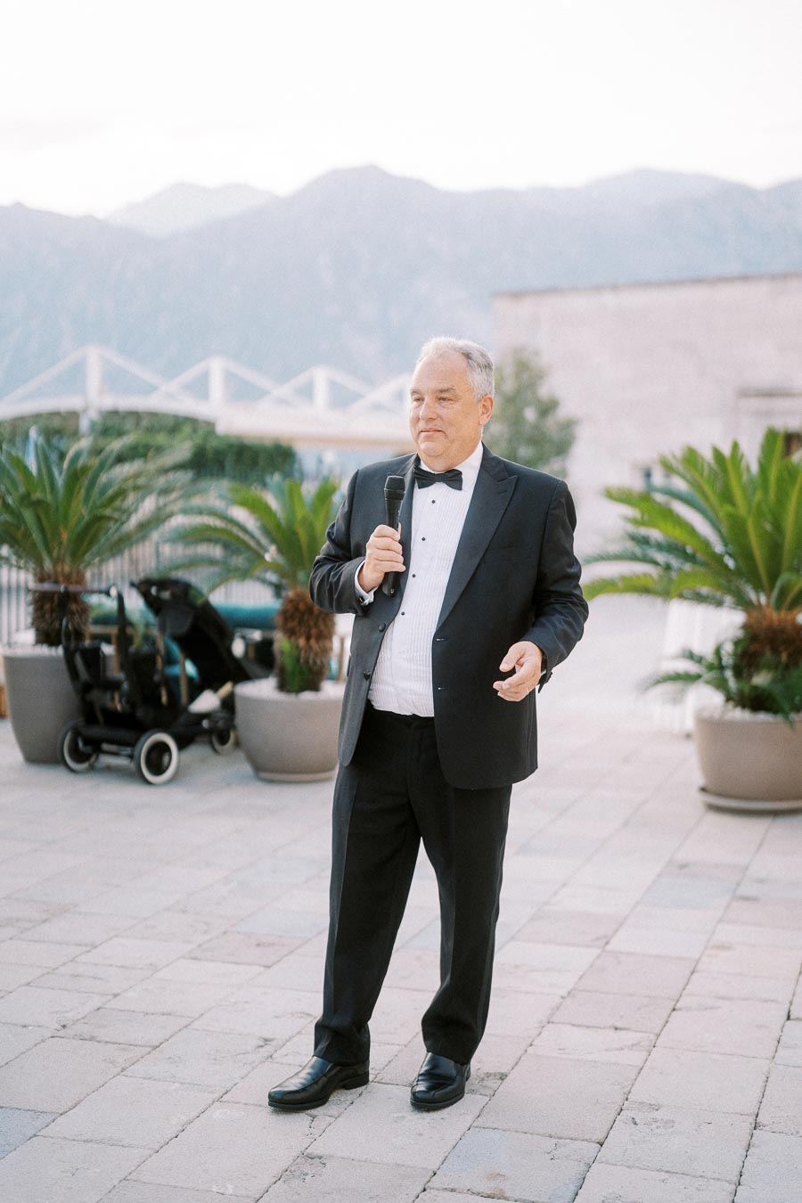 A well-dressed man in a black tuxedo delivers a speech outdoors, holding a microphone. A scenic mountain range and potted plants serve as the backdrop, with a stroller visible beside him.