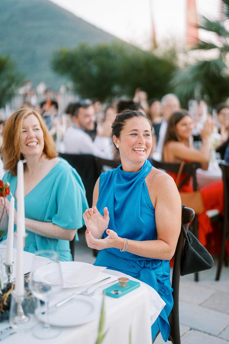 Guests at an outdoor event clapping and smiling during a sunny day, with a scenic mountain backdrop. Women wearing elegant blue dresses seated at a table with white tablecloth and candles, creating a joyful and festive atmosphere.