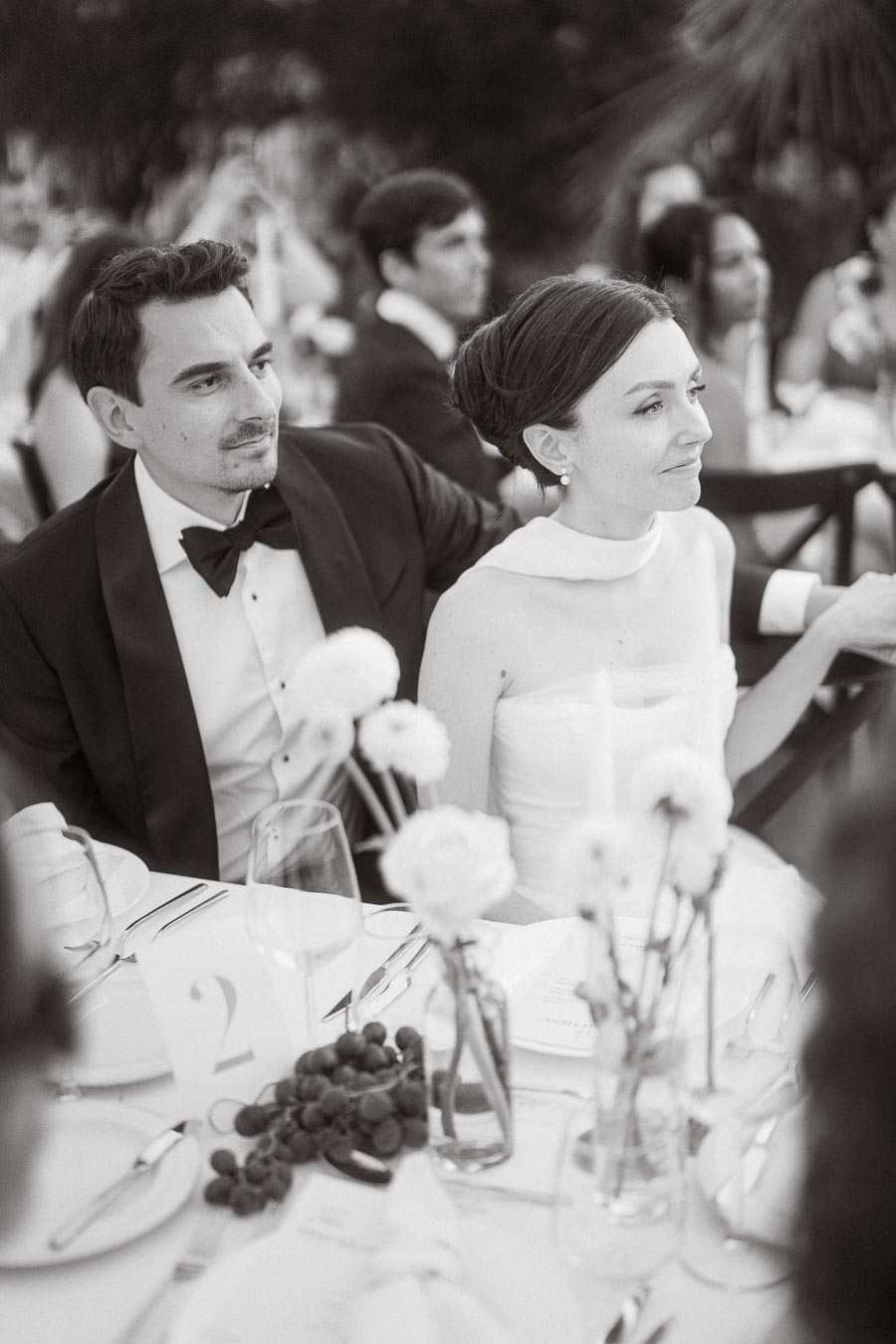 A black and white image of a bride and groom sitting at a wedding reception table, elegantly decorated with flowers and grapes, capturing a moment of joy and elegance.