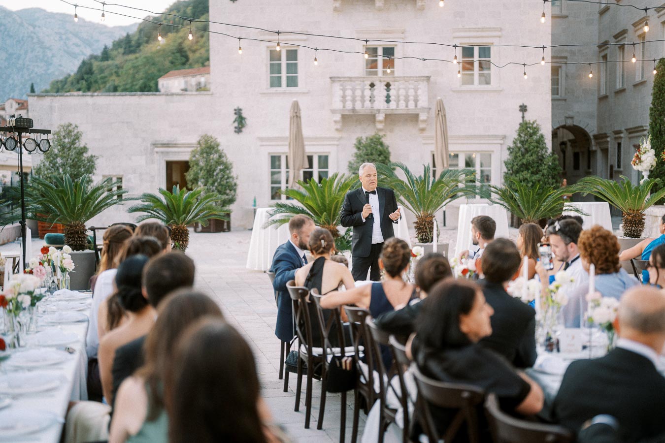 Outdoor wedding reception with a man giving a speech to seated guests, elegant stone building and string lights in the background.
