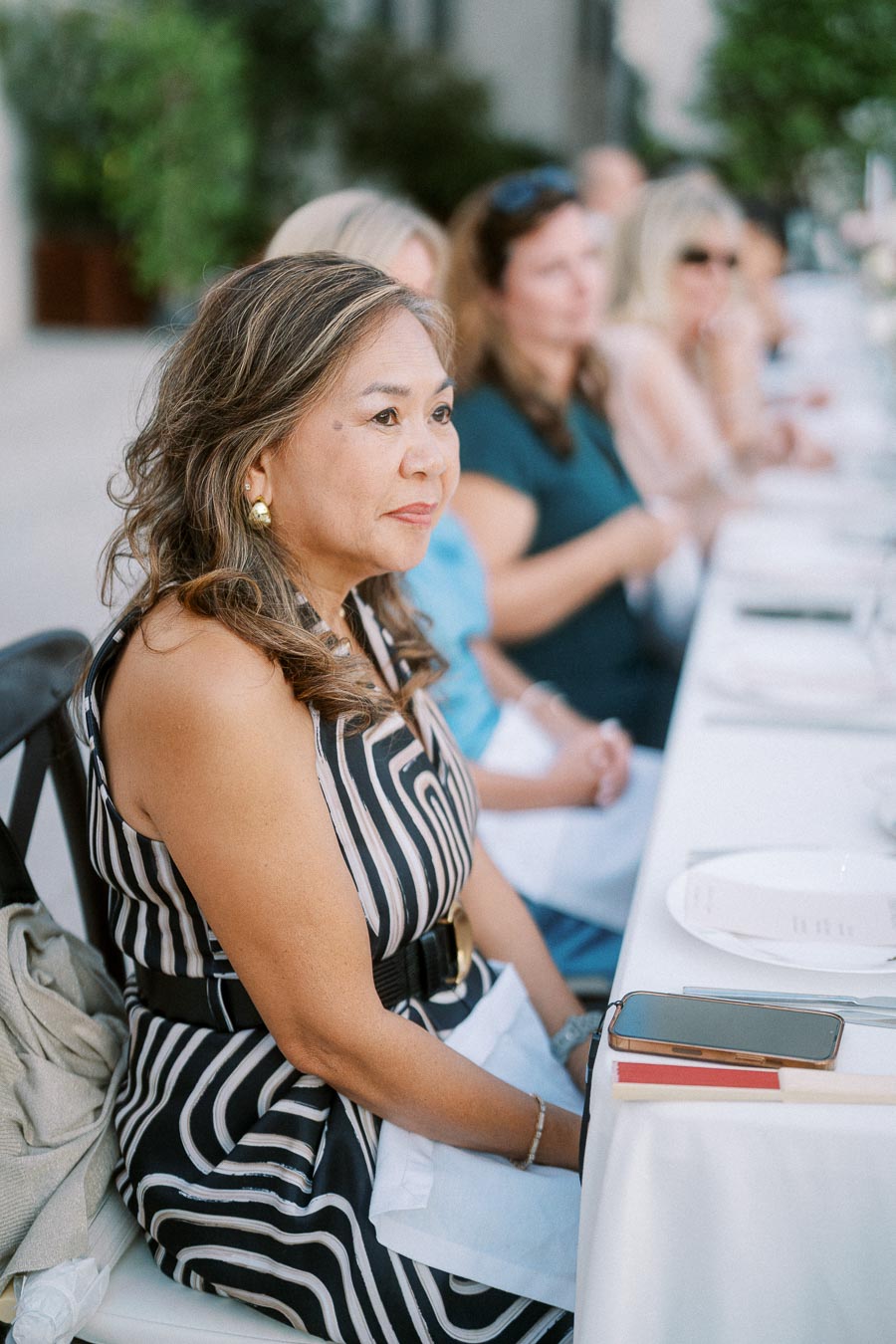 Outdoor dining event with people seated at a long table, featuring a woman in a black and white striped dress in the foreground, attentively listening.