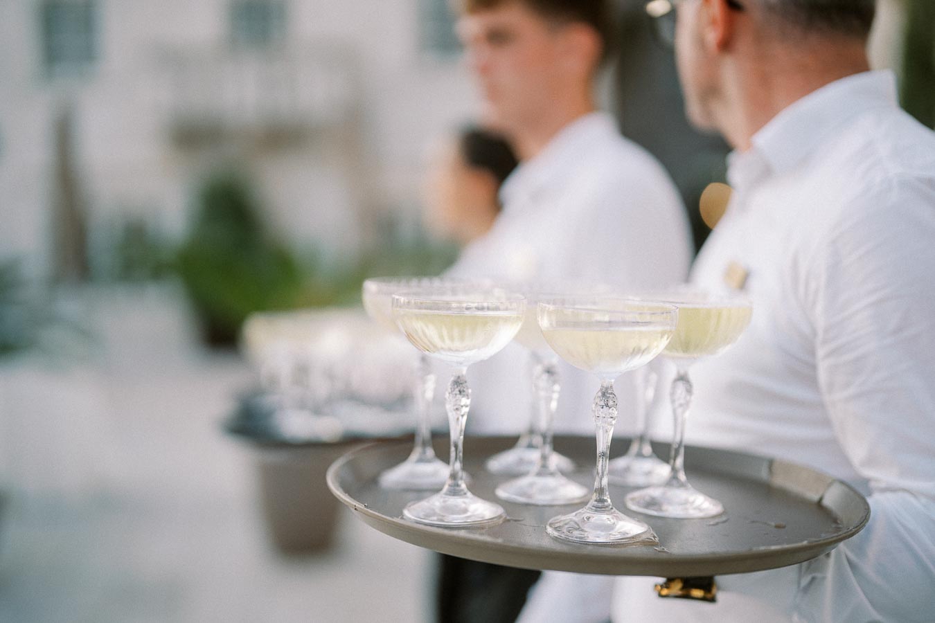 Tray of elegant champagne glasses carried by waitstaff in white uniforms at an outdoor event.