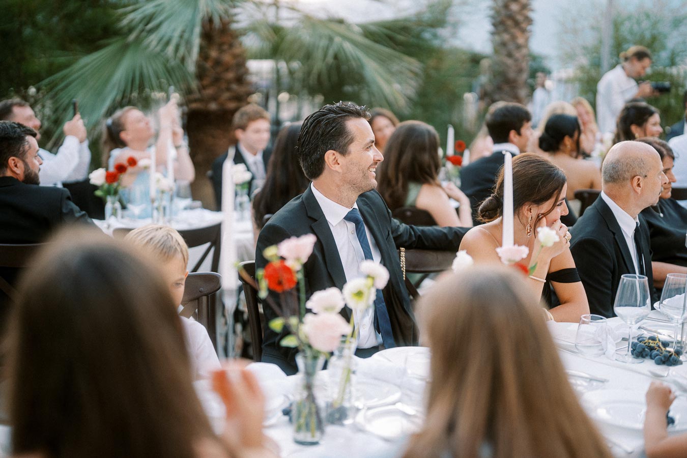 Outdoor wedding reception with guests seated at elegantly decorated tables, surrounded by palm trees and floral centerpieces, creating a festive and elegant atmosphere.
