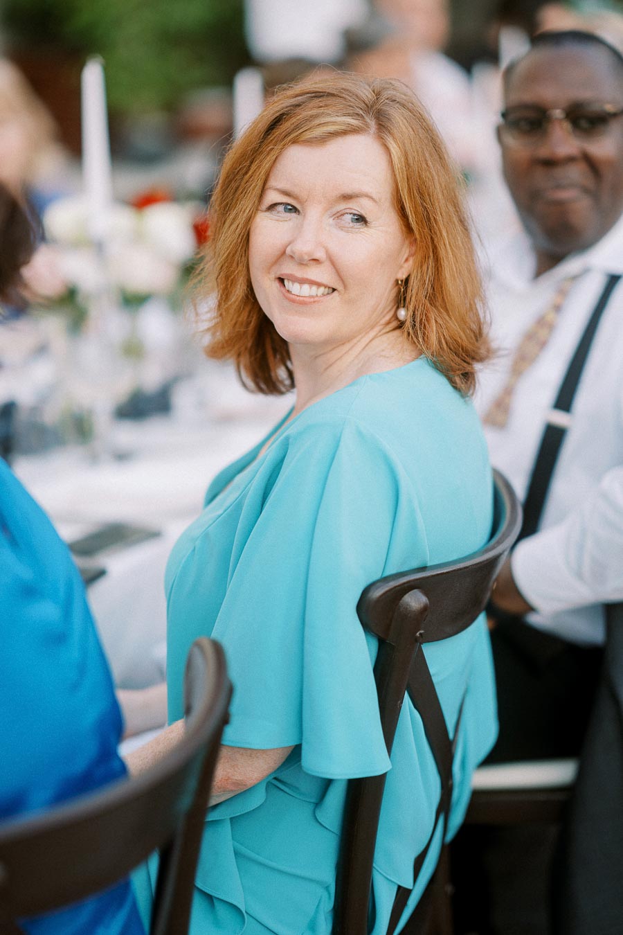 A woman in a light blue dress smiles while sitting at an outdoor event, with blurred guests and white table decorations in the background.