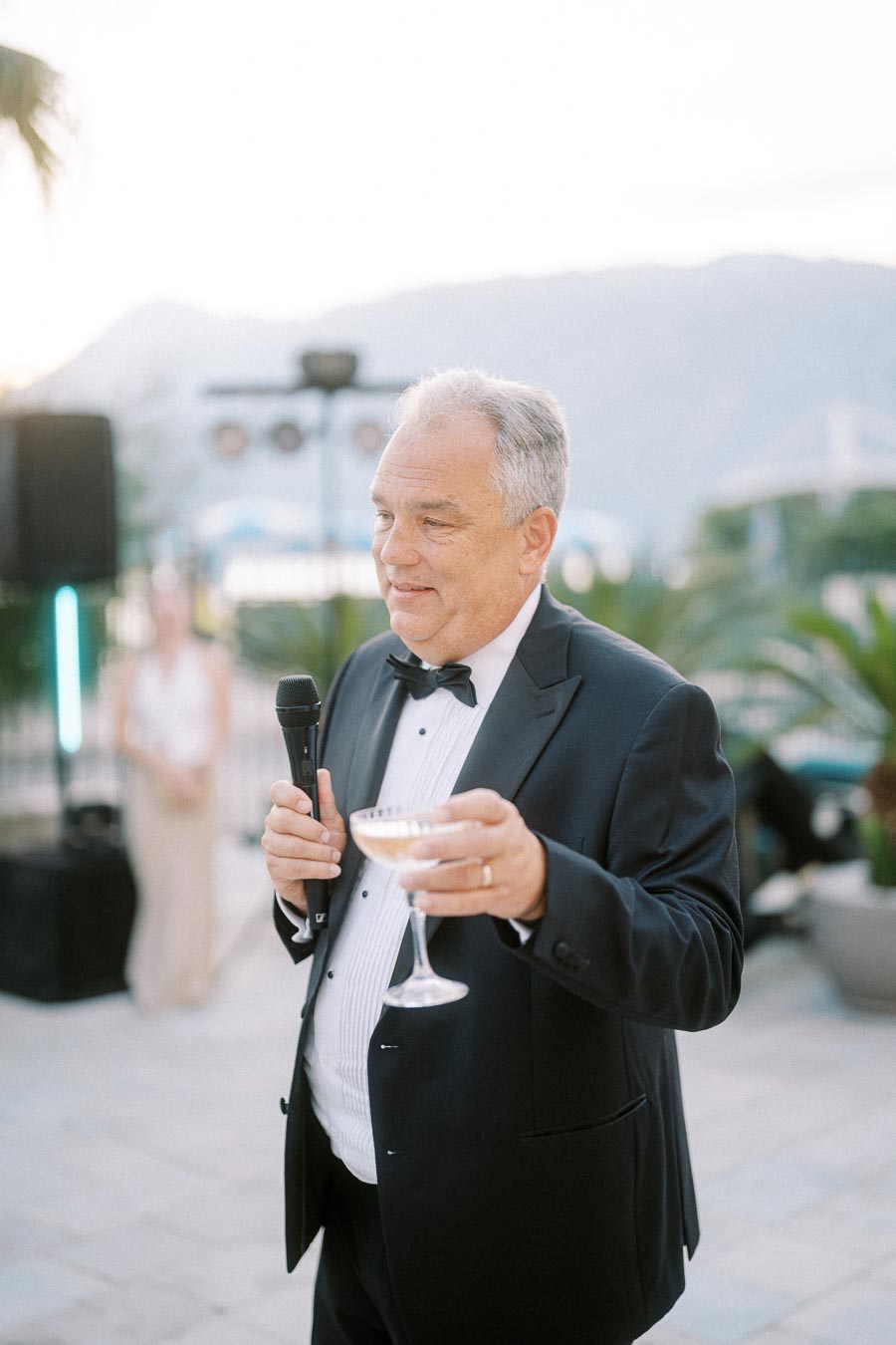 Formal event speaker holding a microphone and a glass, wearing a black tuxedo, with an outdoor background featuring palm trees and mountains.