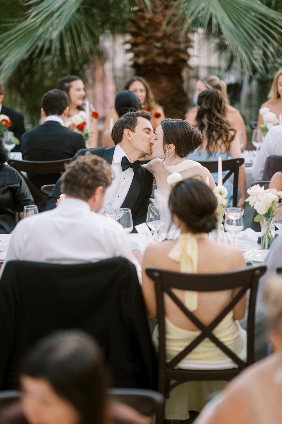 A couple sharing a romantic kiss at an outdoor wedding reception, surrounded by elegant decor and guests seated at tables under palm trees.