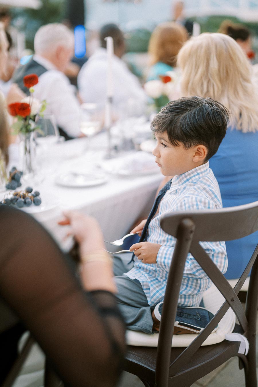 A young boy in a blue checkered shirt sits attentively at a formal dining table during a festive outdoor event, surrounded by guests and floral centerpieces.