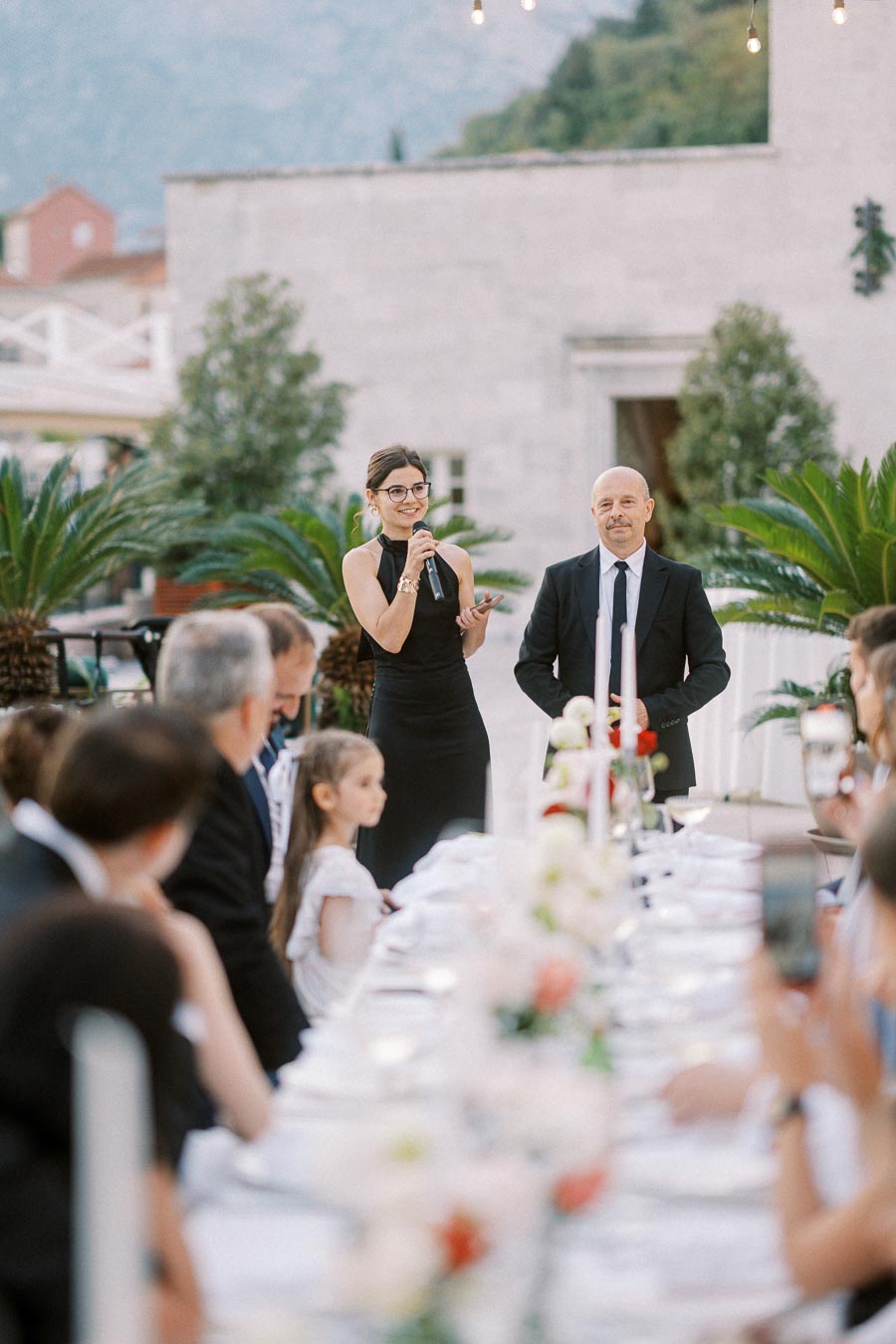 A woman in a black dress speaking into a microphone at an outdoor formal gathering, with people seated around a long table decorated with flowers and candles.