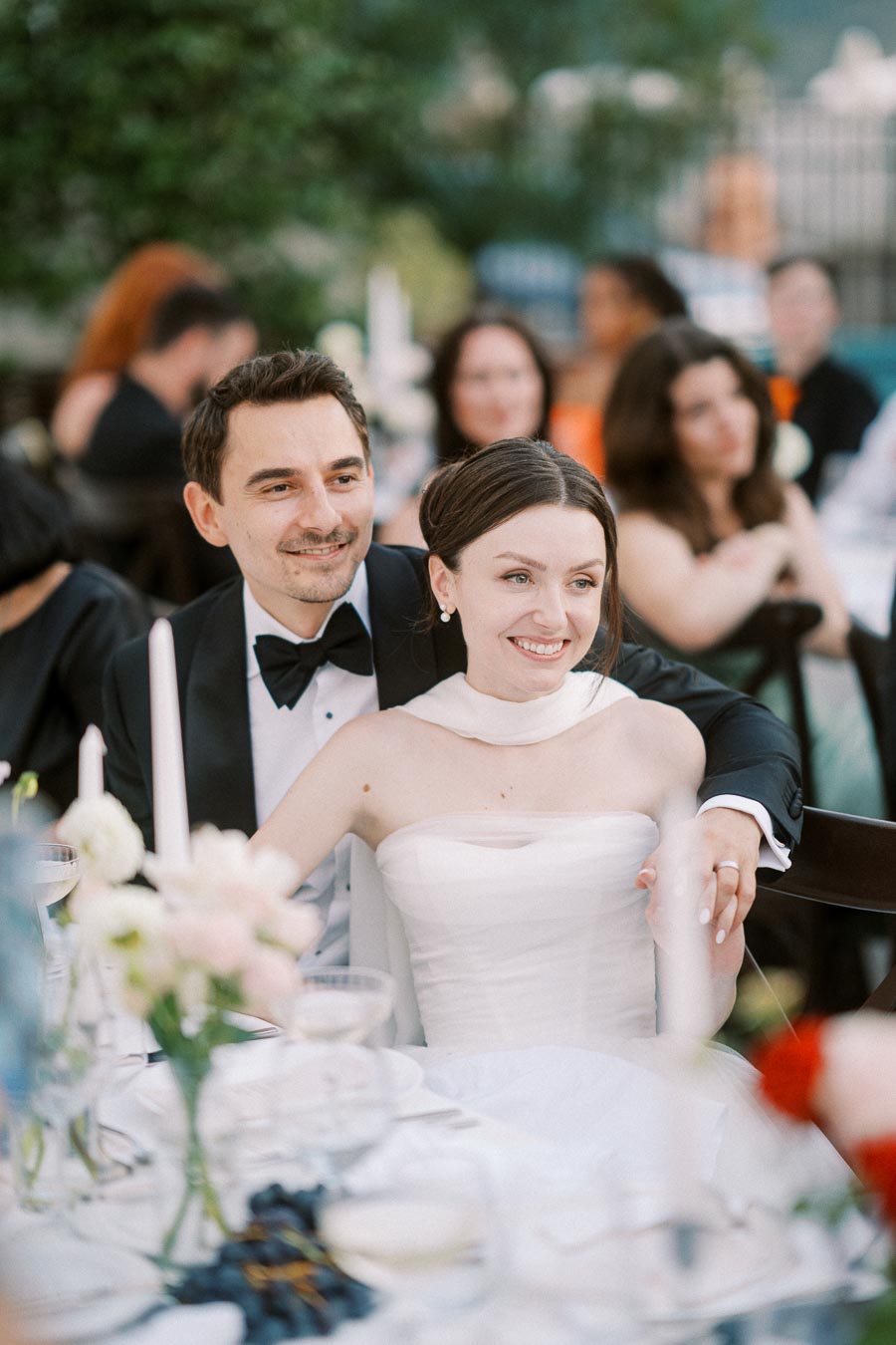 Elegant bride and groom smiling during outdoor wedding reception, with guests in the background and a beautifully set table with flowers and candles.