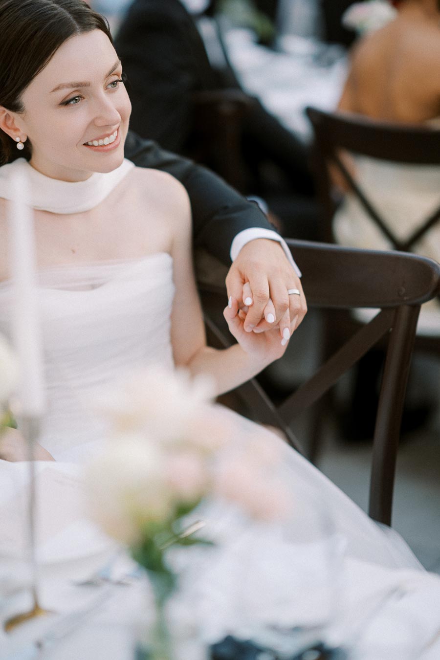 A smiling bride in a white wedding dress holds hands with her partner, showcasing a close-up of their wedding rings during a reception.
