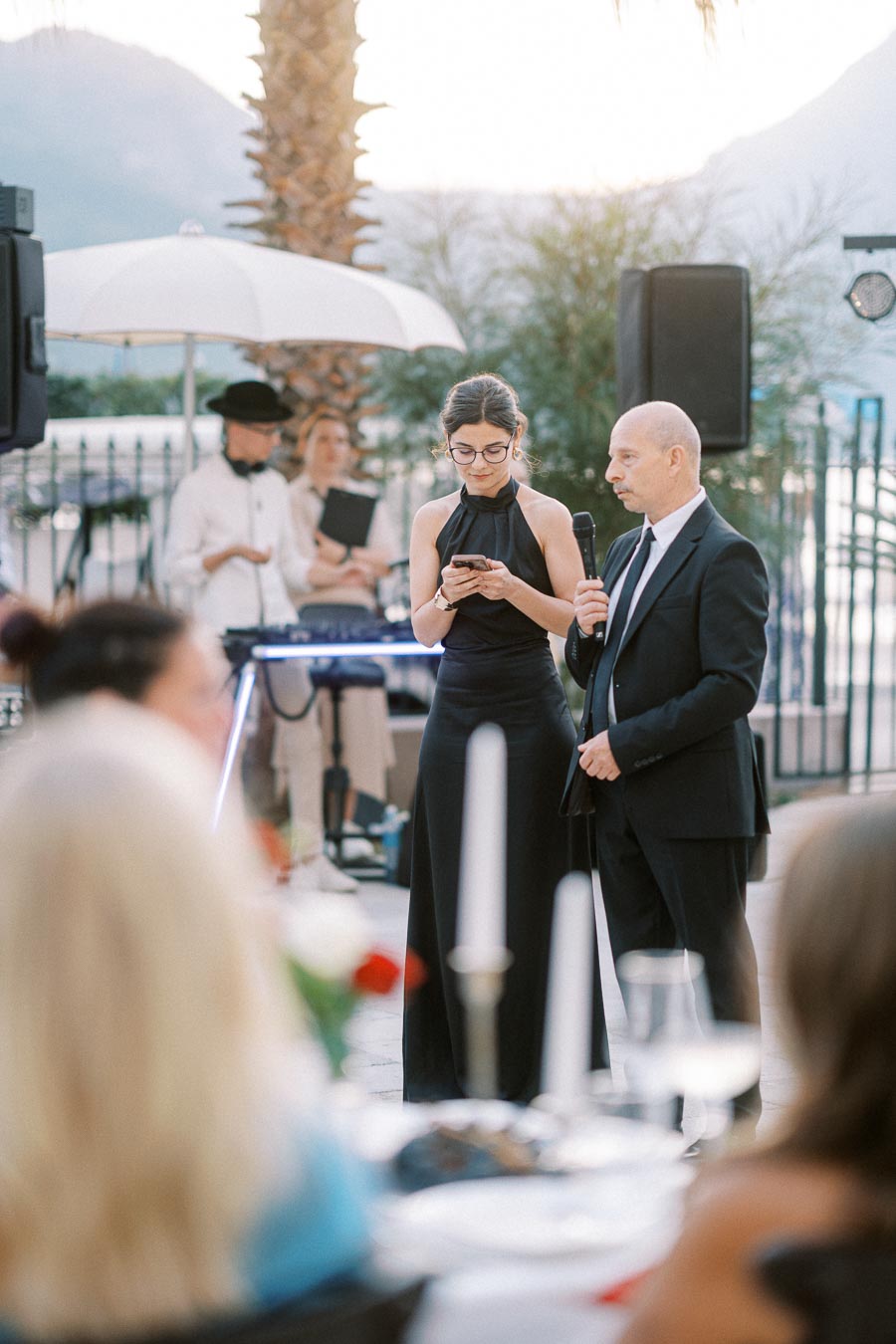 Wedding reception speech with two elegantly dressed individuals; a woman in a black dress checks notes on her phone while standing beside a man in a suit holding a microphone, under an outdoor setting with ambient lighting and a musician in the background.